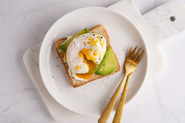 Savoury breakfast - A toast bread with cream cheese, avocado and poached egg on a white plate on marble tray