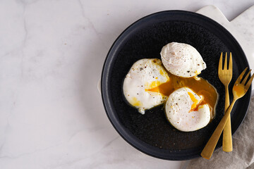 Three poached eggs with egg yolk on a black plate on a marble board and golden forks
