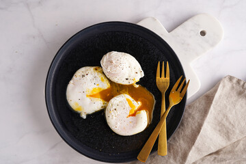 Three poached eggs with egg yolk on a black plate on a marble board and golden forks