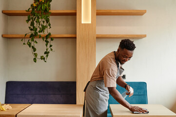 Medium long shot of young African American man wearing apron working in small cafe preparing tables...