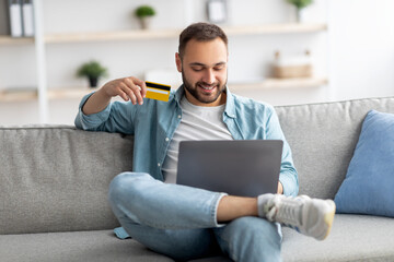 Happy young Caucasian guy with laptop and credit card sitting on sofa at home, shopping online in web store