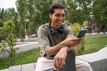 joyful african american skater sitting on border bench and messaging on smartphone.