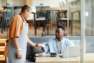 Young Black man having lunch in modern cafe sitting at table paying for order with bank card