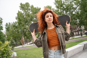 young curly woman looking at camera while holding skateboard in city park.