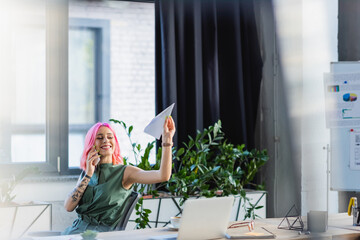 positive businesswoman with pink hair talking on smartphone while throwing paper plane.