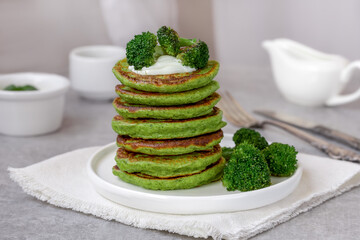 Fluffy broccoli green pancakes served with yoghurt sauce and fried broccoli florets in white plate. Vegetable fritters. Healthy Breakfast. Selective focus, gray concrete background.