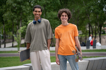 joyful interracial friends with skateboards smiling at camera outdoors.