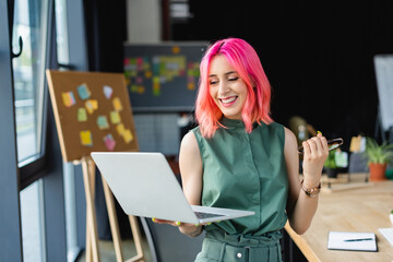 joyful businesswoman with pink hair holding laptop and eyeglasses in office.