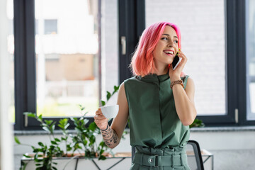 cheerful businesswoman with pink hair and piercing holding cup of coffee while talking on smartphone in office.