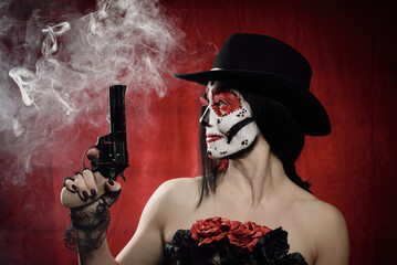 beautiful woman in skeleton make-up holds a revolver, she is wearing a black hat. White smoke over...