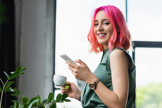 cheerful businesswoman with pink hair and piercing holding cup of coffee and smartphone.