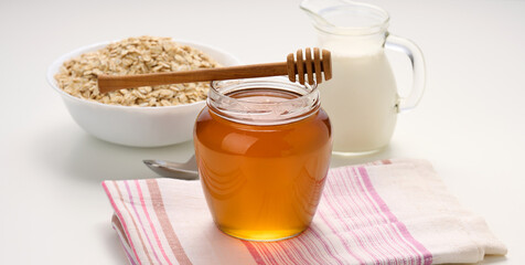 honey in a glass transparent jar and a wooden stick on a white table, behind a decanter with milk