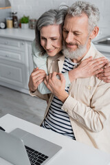Mature couple hugging near laptop in kitchen at home.