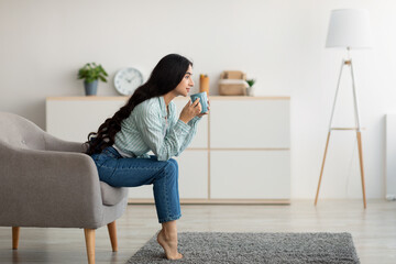Side view of young Indian woman sitting in armchair, sipping hot coffee, enjoying lazy relaxing day...