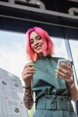 low angle view of pleased businesswoman with pink hair holding smartphone.