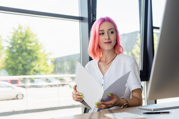 pierced businesswoman with pink hair holding folder and looking at computer monitor.