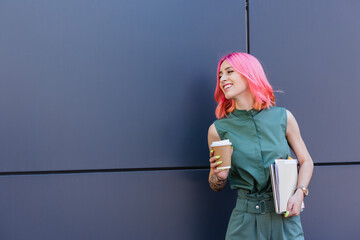 tattooed and smiling businesswoman with pink hair holding folder and coffee to go outside.