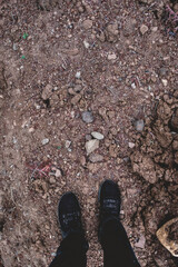 Feet with black sneakers and and black jeans standing over dirt soil with stones (texture, background)