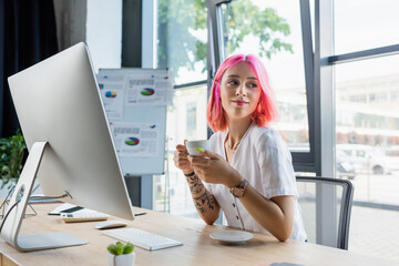 young pierced manager with pink hair holding cup of coffee in office.