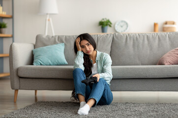 Obraz premium Full length of unhappy Indian woman sitting on floor with joystick, losing online video game, feeling stressed at home