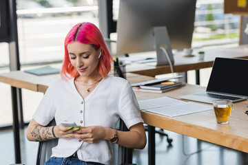smiling young businesswoman with pink hair and earphone using smartphone in office.