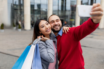 Loving young diverse couple with gift bags taking selfie near huge shopping centre