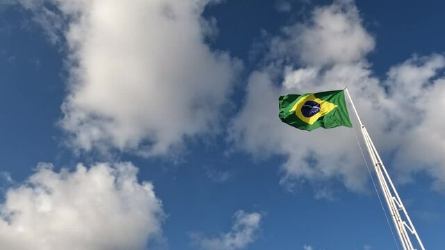 Huge flag of brazil waving freely under a sky with some white clouds.