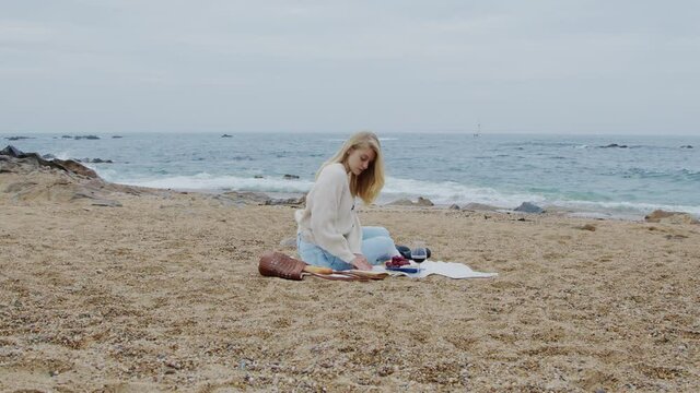 Blonde woman is sitting on blanket, enjoying her picnic on ocean beach, reading interesting book, taking pleasure of her lifestyle, young and successful, Zoom In, Slow motion.