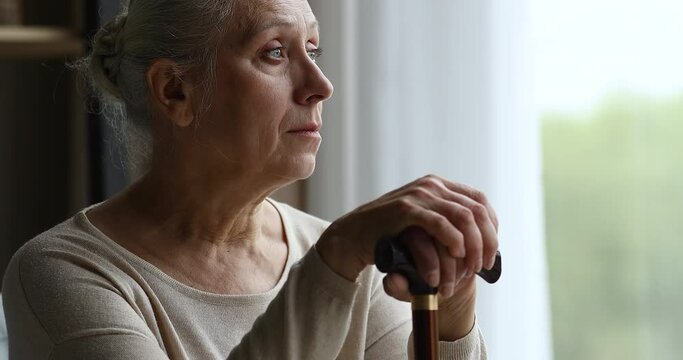 Cropped close up of depressed hoary older adult female living with physical disability sit by window alone hold stick. Retired old woman with walking difficulties look at distance feel weak hopeless