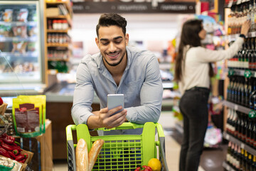 Arab Male Customer Using Cellphone For Grocery Shopping In Supermarket - obrazy, fototapety, plakaty