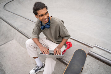 Obraz premium high angle view of cheerful african american student using laptop on border near skateboard.
