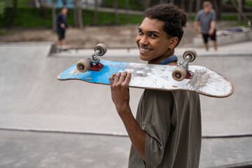young african american man smiling at camera while holding skateboard on shoulder.