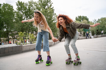excited women holding hands while rollerblading in park. - obrazy, fototapety, plakaty