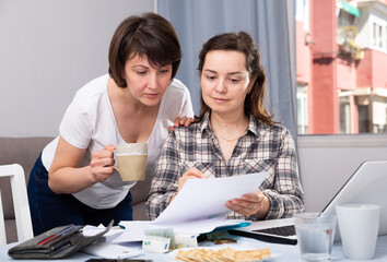 Two smiling young females looking financial documents with laptop in home interior. High quality photo