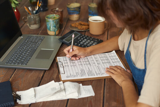 Young woman making notes while working with financial papers in front of laptop by wooden table in workshop