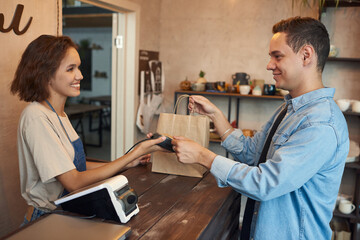 Young female shop assistant of craft store passing paperbags with purchase to male buyer paying by credit card