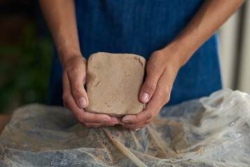 Hands of young creative woman holding large piece of clay over workplace while going to knead or roll it