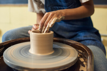 Hands of young female potter sitting by rotating pottery wheel while working over new collection of earthenware