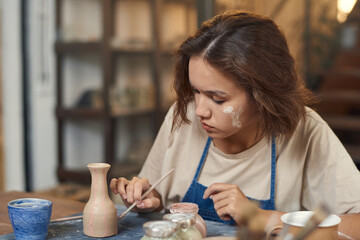 Young creative woman with paintbrush painting self-made jug or pot while sitting by workplace in front of camera