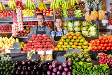 Fototapeta na wymiar Young man and woman sellers in aprons standing near counter with a fresh vegetables and fruits on market