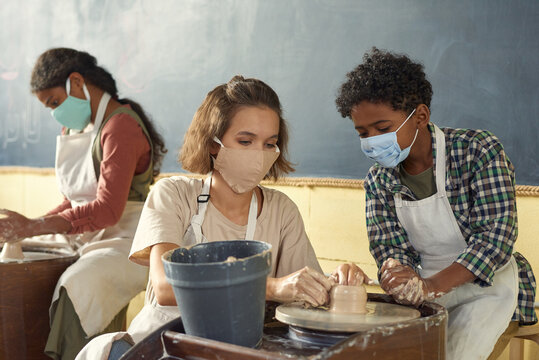 Teacher and schoolboy in protective masks sitting by pottery wheel while boy learning how to sculpt clay pots and other earthenware