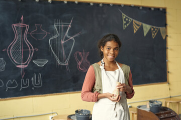 Happy cute schoolgirl with handmade clay pot standing by blackboard with sketches of earthenware in...
