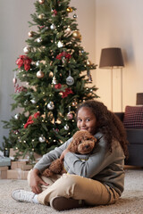 Happy girl with cute pet looking at you while sitting on the floor of living-room with decorated xmas tree on background