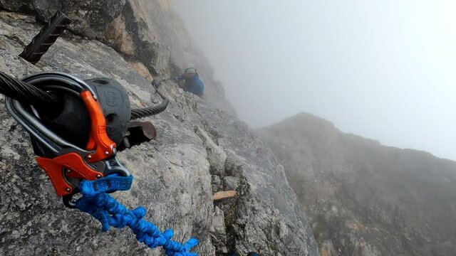 PoV shot of a climber looking down at another men climbing up a very steep cliff. They are in the clouds, so it looks very mystical and dangerous.