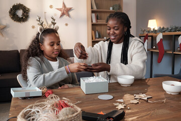 Cute African girl in headband helping her mother with packing of xmas gifts while sitting by table in living-room