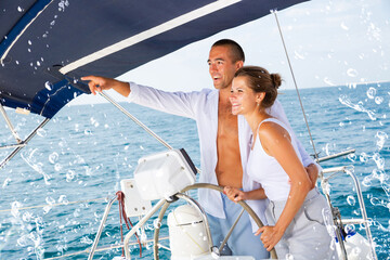 Smiling man pointing to something on ocean horizon to his wife while standing on yacht wheel enjoying summer sea vacation