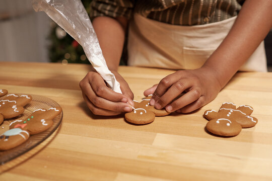 Hands of African girl decorating homemade gingerbread cookies with white glaze while standing by wooden table