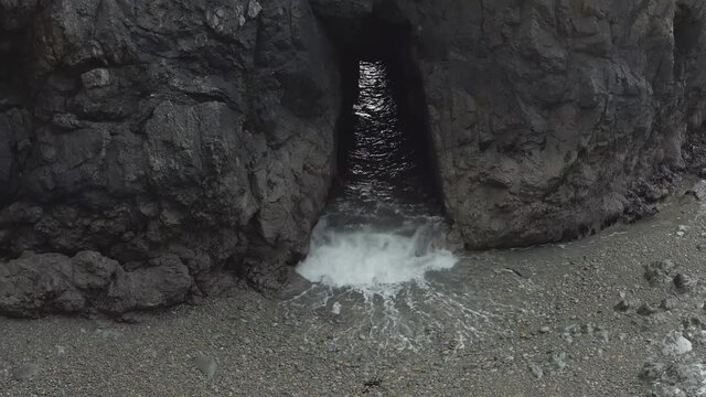 Tunnel in rocky shore islet allows grey ocean waves to surge through