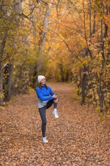 Outdoor pursuit. Caucasian fit woman in blue sportswear and a knit cap warning up her knees before jogging in the forest in autumn, vertical photo, selective focus