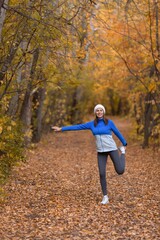 Caucasian attractive smiling woman in blue sportswear and a knit cap warning up her legs before jogging in the forest in autumn, vertical photo, selective focus